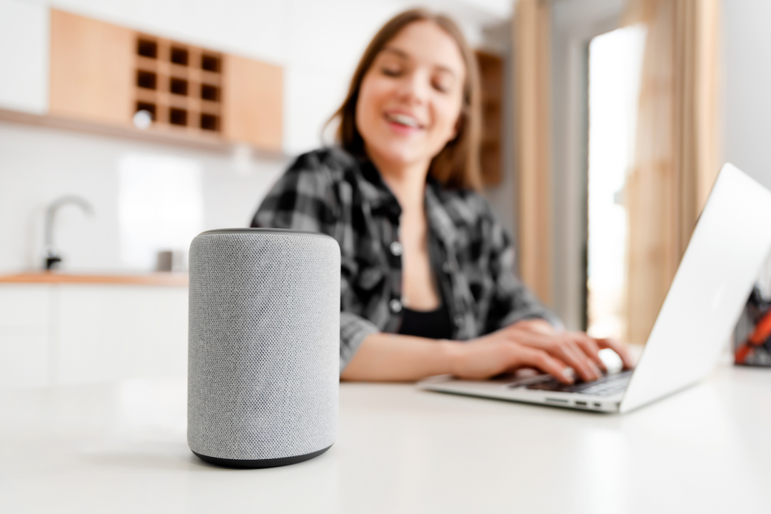 A modern living room featuring a voice assistant device (smart speaker) on a table. A young professional is speaking to the device while multitasking.
